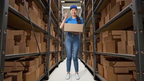 full body of asian female courier in blue uniform showing gesture peace and smiling while delivering a carton in warehouse