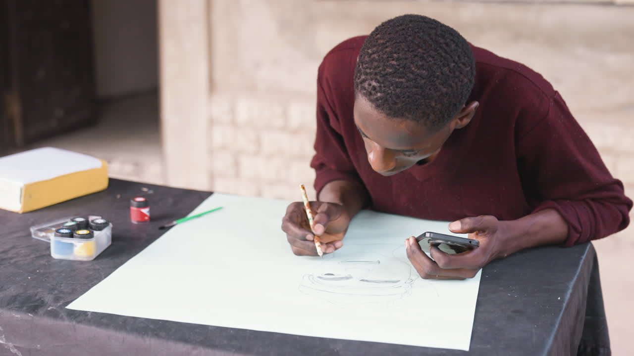 Homme noir peignant en extérieur sur une table, une feuille de papier pâle et des pots de peinture à proximité, pull marron, fond de porche et fenêtre, lents coups de pinceau de finition et humeur calme et pensive, main ferme composant