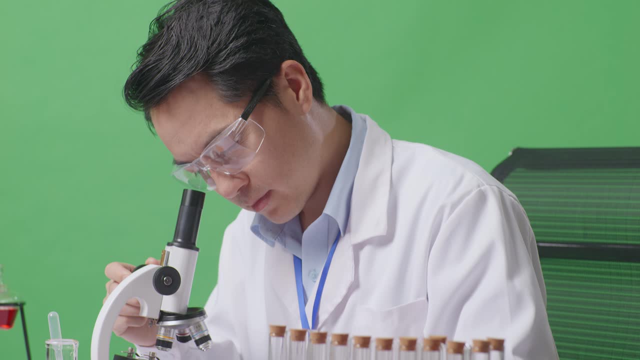 Close Up Of Side View Of Asian Man Scientist Thinking, Looking Around, And Raising Index Finger While Looking Through A Microscope On The Table With Test Tube In The Green Screen Background Laboratory