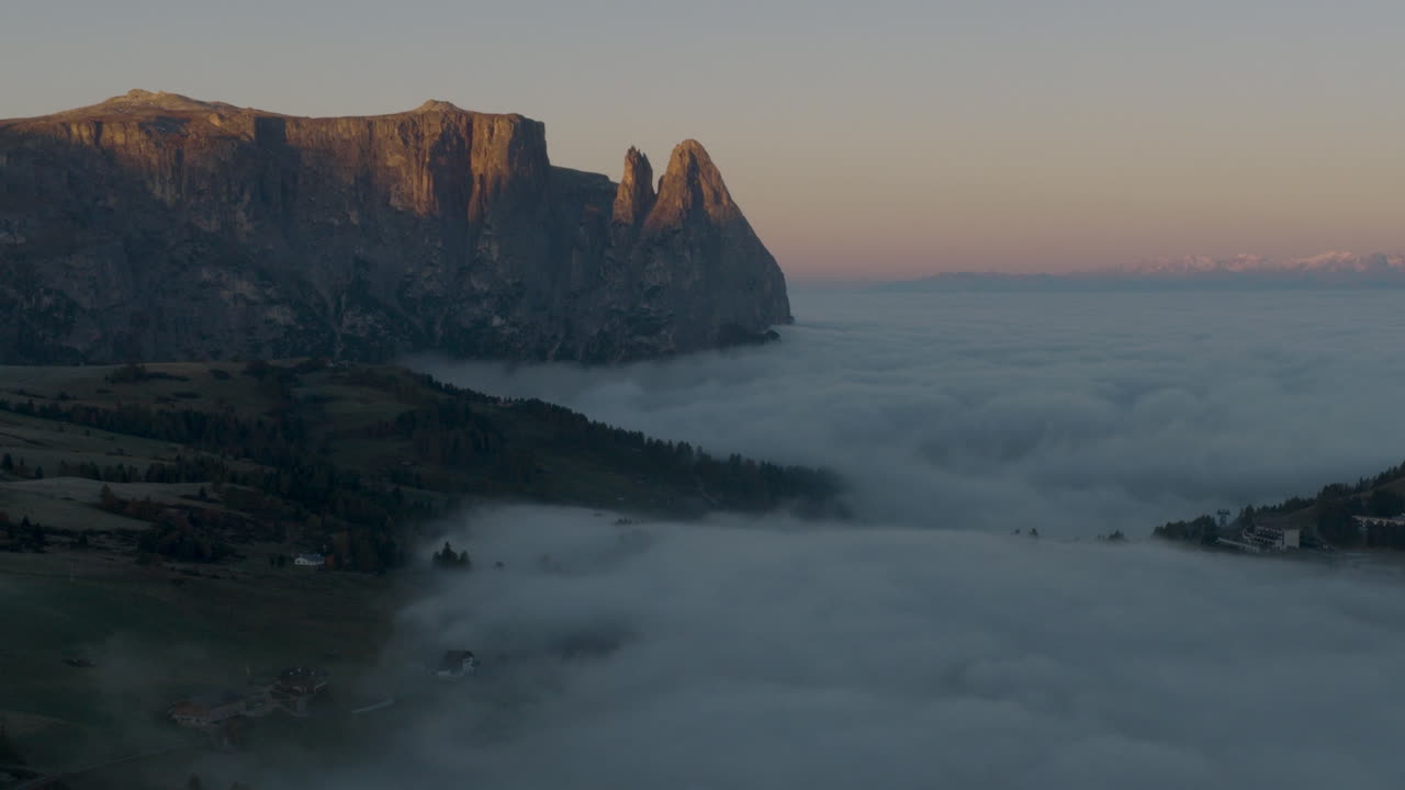 Cinematic aerial view of frosty Alpe di Siusi meadows and scattered mountain huts at dawn, mist drifting across rolling hills beneath the dramatic Sassolungo peaks in the Dolomites, Italian Alps