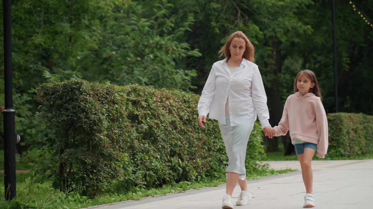 Kind en moeder wandelen onder bladerrijke bomen. Moeder en dochter voeren een informeel gesprek tijdens een zonnige parkwandeling. Een sterke familieband wordt getoond terwijl ze samen genieten van een zonnige middag buiten.