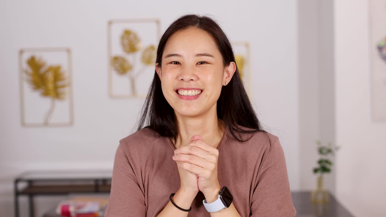 Young woman smiles, clasps hands in gratitude, warmly lit home interior, static camera, cheerful mood