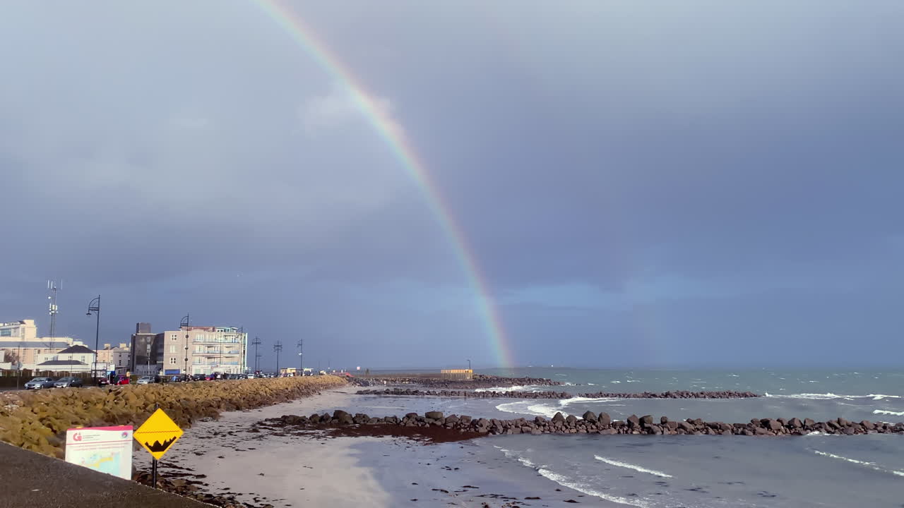 Handheld shot of Salthill Promenade as waves roll in, with a vibrant rainbow arching over the seascape
