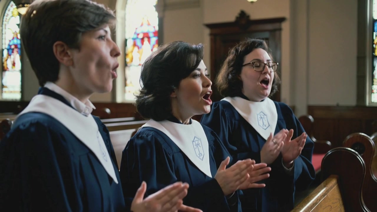Trio di coro che canta seguendo l'indicazione del direttore, applaudendo al banco della chiesa, con tonache blu navy e colletti bianchi.