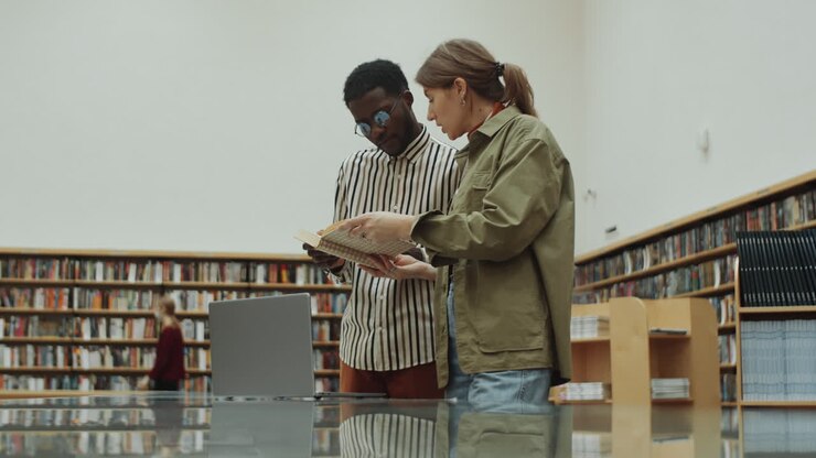Uomo e donna multietnici che leggono un libro e usano un laptop in biblioteca.