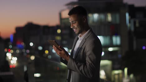 Video-of-smiling-biracial-businessman-using-tablet-at-night-on-office-roof-terrace