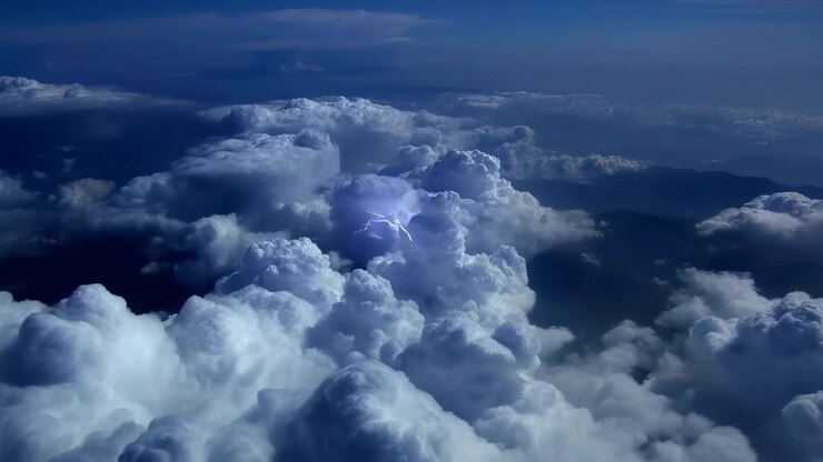 Bright internal lightning briefly illuminates textured cloud towers while the view glides above layered storm formations, with cool blue light, deep shadows, and distant cloud bands.