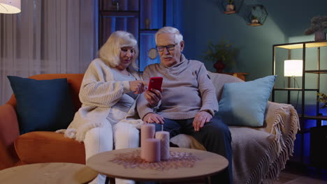 senior couple grandfather grandmother making online shopping on mobile phone sitting at home sofa