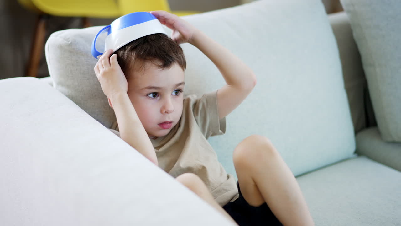 Child plays with bowl on head at home. A young boy sits on a couch wearing a bowl as a hat, looking pensive while playing indoors