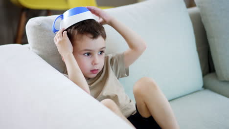child plays with bowl on head at home. a young boy sits on a couch wearing a bowl as a hat, looking pensive while playing indoors