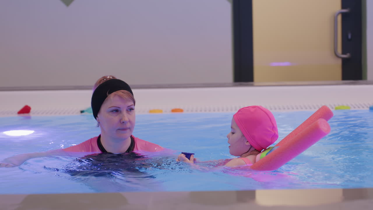 Kid scooping water inside pool with toy cup during playful lesson tiny hands pouring supervision nearby early learning motor skill practice clean blue surface joyful movement gentle guidance