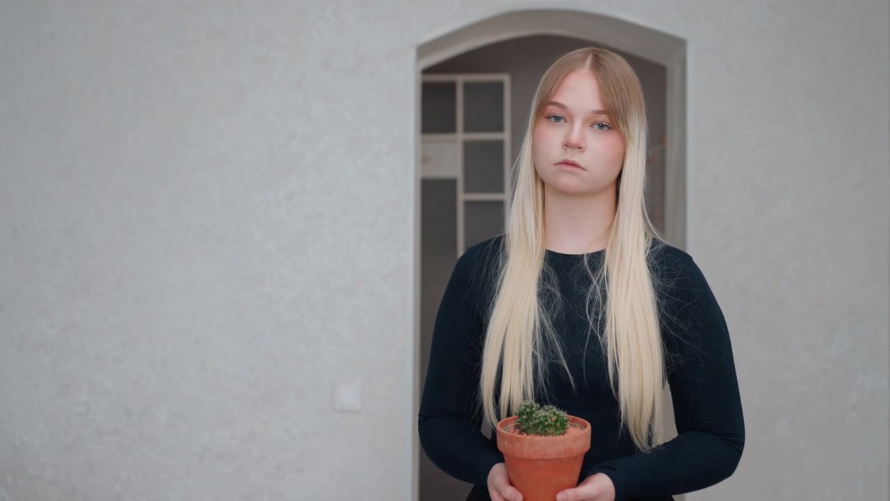 Nuori valkoihoinen nainen pitelee ruukkukasvia tyhjässä uudessa asunnossa, uusi asunnonomistaja purkaa laatikoita ja miettii kasvin sijoittelua kaarevan oviaukon lähelle. Minimalistinen, neutraali sisustus, pehmeä valo, pitkät varjot.