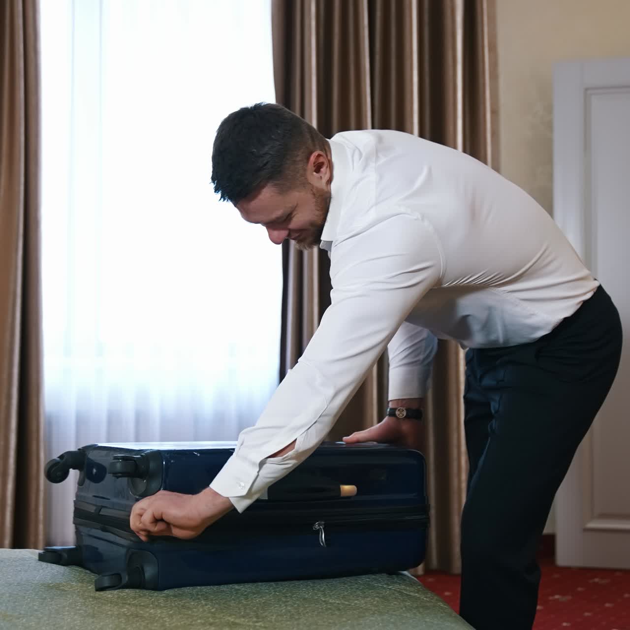 Happy businessman leaving hotel room. Young man in white shirt and trousers closes his luggage. Traveller finishes packing stuff and ready to leave hotel