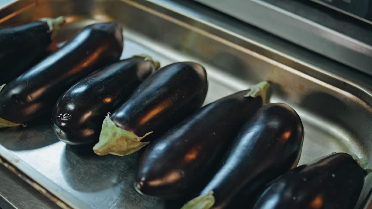 Woman hand putting eggplants on metal tray at cafeteria kitchen closeup