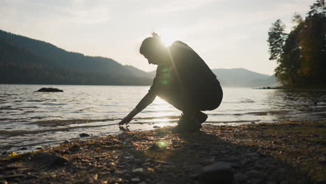 lady puts fingers down into lake at sunset. woman squats by water alone discovering calm nature in autumn evening. perfect moment to enjoy solitude