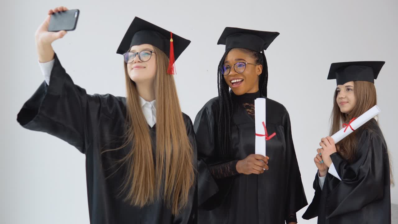 tres jóvenes graduadas felices de diferentes nacionalidades se unen y se toman una selfie con diplomas en las manos. vista de cerca sobre fondo blanco