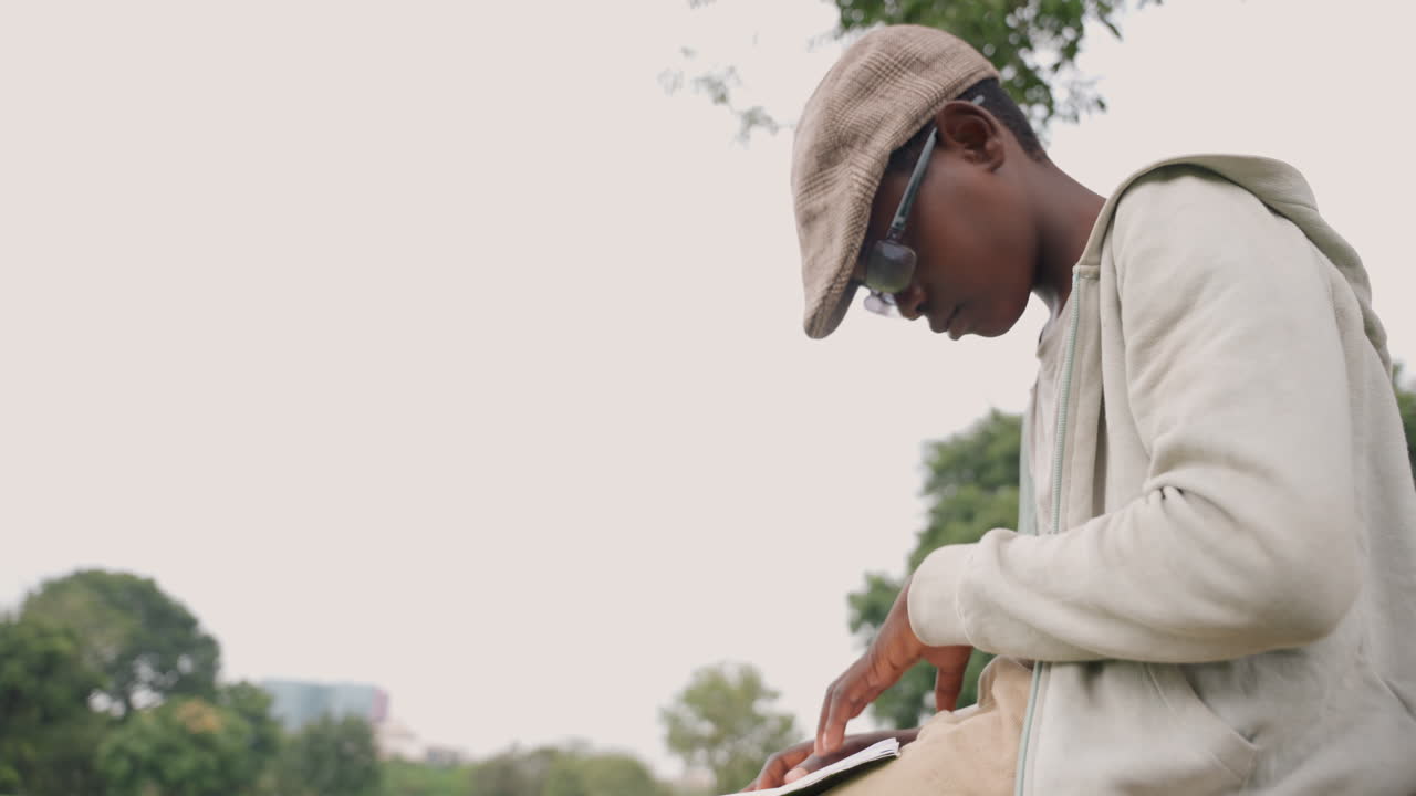 Closeup side profile son outdoors reading and typing on phone, casual cap and hoodie, soft light, urban park trees in background, focused expression conveying reflection and quiet study