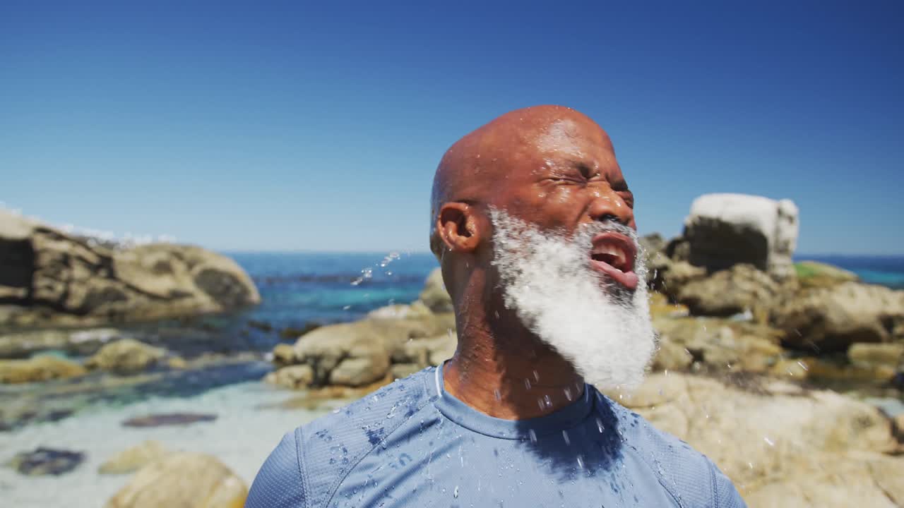 hombre afroamericano mayor ejercitando verter agua en la cara en las rocas junto al mar