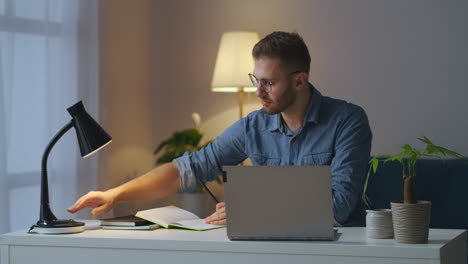 Hübscher-Männlicher-Student-Lernt-Online-Am-Abend,-Schreibt-In-Ein-Notizbuch-Und-Sucht-Informationen-Auf-Dem-Laptop,-Sitzt-Am-Tisch-Im-Wohnzimmer,-Porträt-Eines-Mannes-Im-Innenbereich