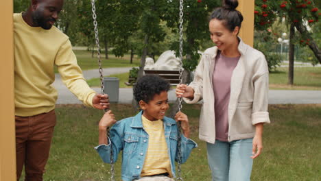 little african american boy swinging on swing in park with mother and dad