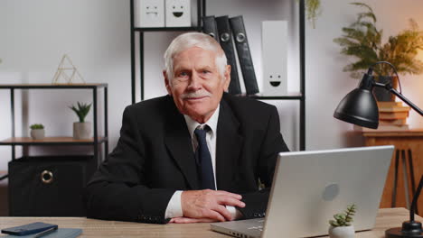 portrait of senior businessman in suit sitting at table with laptop smiling at camera at home office