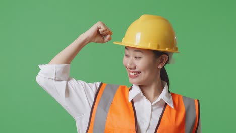 close up of asian female engineer with safety helmet flexing her bicep and smiling to camera while standing in the green screen background studio