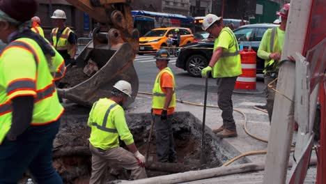a street level shot of workers digging a trench on fifth avenue in nyc on a cloudy day. two men are in the hole, as others stand around, all in neon green safety vests. the camera is stationary.