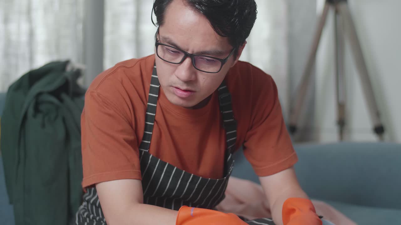 primer plano de una ama de llaves asiática con un delantal sentada en un sofá y con dolor de cabeza debido a estar cansada limpiando la habitación desordenada