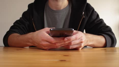 Junger-Mann-Benutzt-Dunkelgraues-Smartphone-Und-Schreibt-SMS,-Sitzt-Auf-Einem-Holztisch