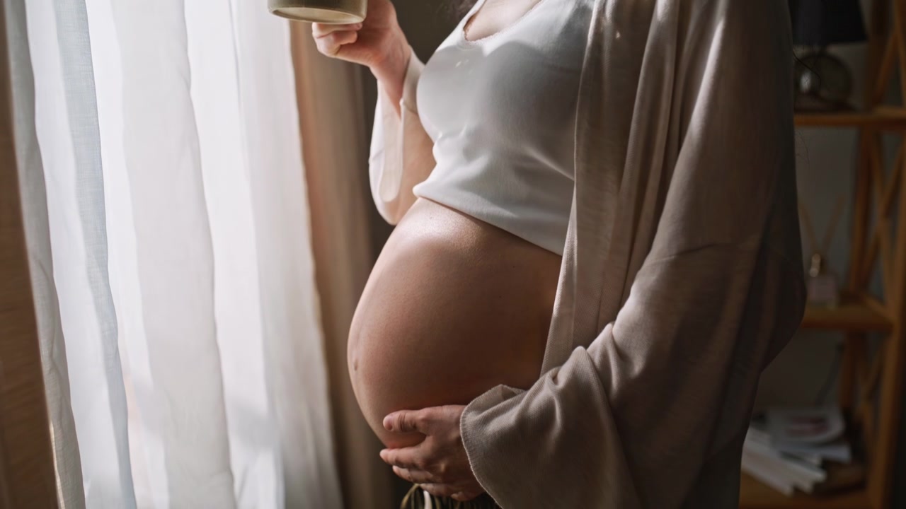 Primo piano di mani che accarezzano la pancia di una donna incinta vicino a una finestra. Futura mamma che beve caffè
