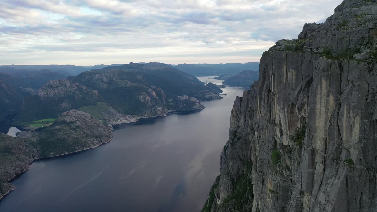 Aerial view of massive cliff towering above winding fjord with still blue water. Rugged mountains and rocky landscape stretch into distance under overcast sky, creating dramatic natural scene
