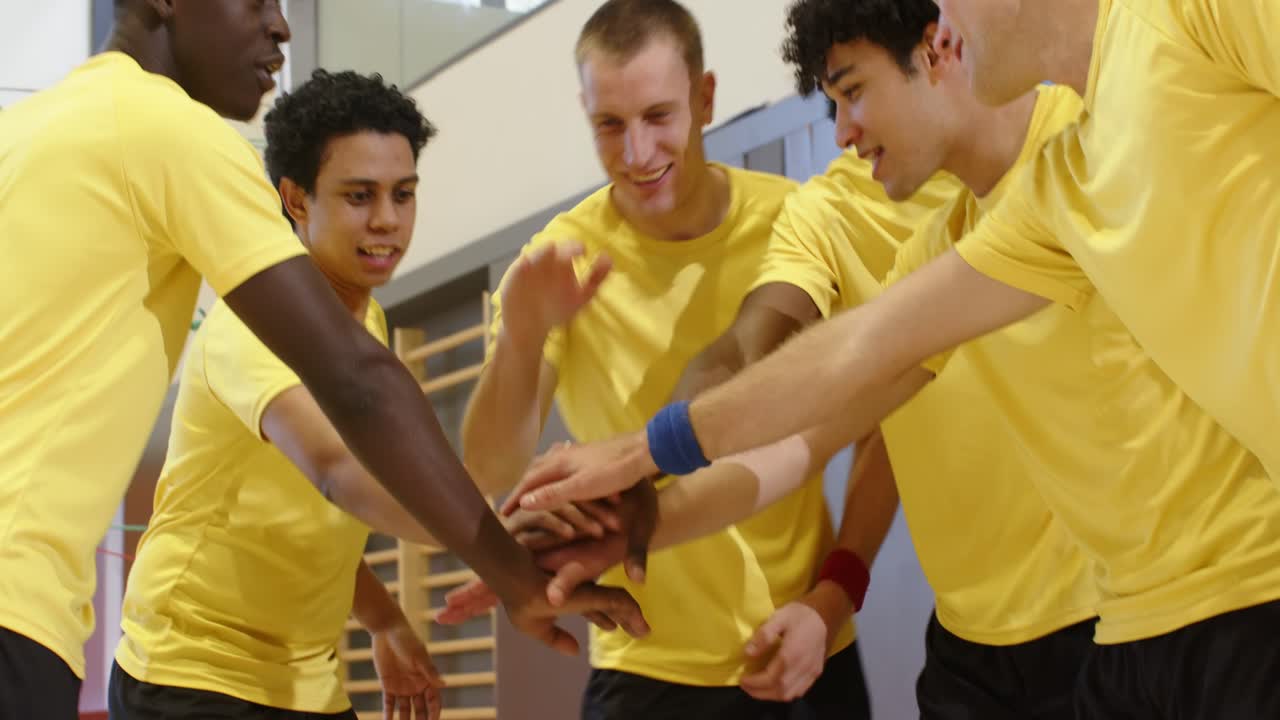 Squadra maschile eterogenea seduta in panchina in palestra che osserva il gioco, applaude, si alza in piedi dopo un'azione chiave e unisce le mani.