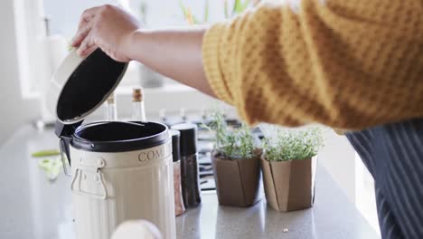 Midsection-of-african-american-woman-in-apron-composting-vegetable-waste-in-kitchen,-slow-motion