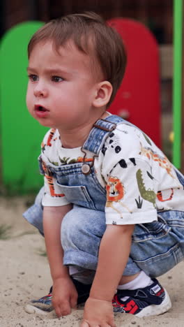 lovely baby boy sits deepening his hands into the sand. little kid looks ahead thoughtfully. vertical video