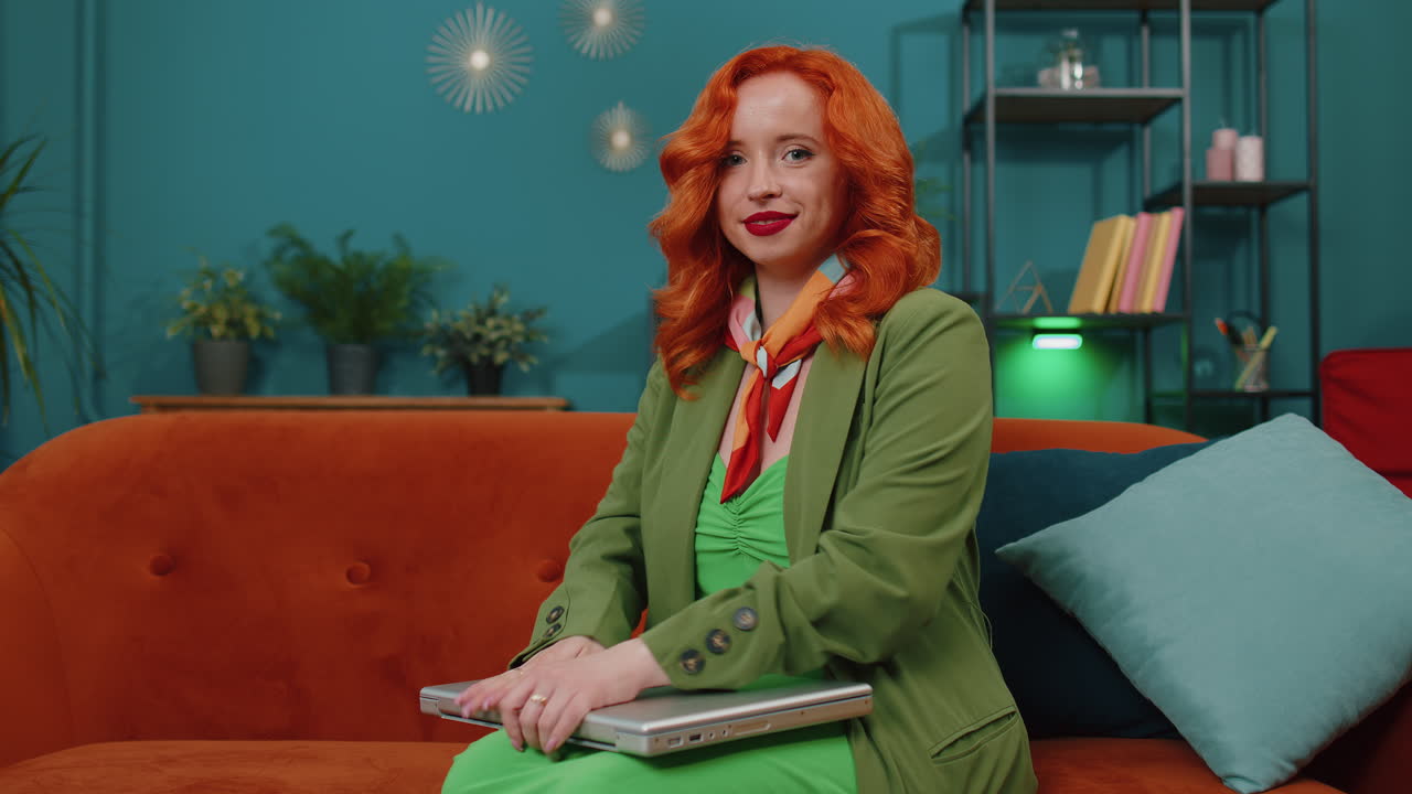 рыжая молодая женщина, сидящая на диване, закрывает ноутбук после окончания работы в гостиной дома