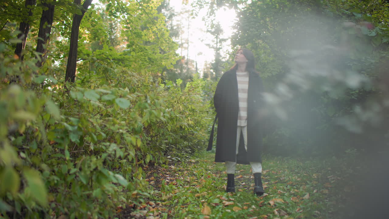 Cozy young girl strolling leisurely along lush forest path, hands on jacket, gazing around with thoughtful calm expression surrounded by green foliage and fallen leaves under soft natural sunlight