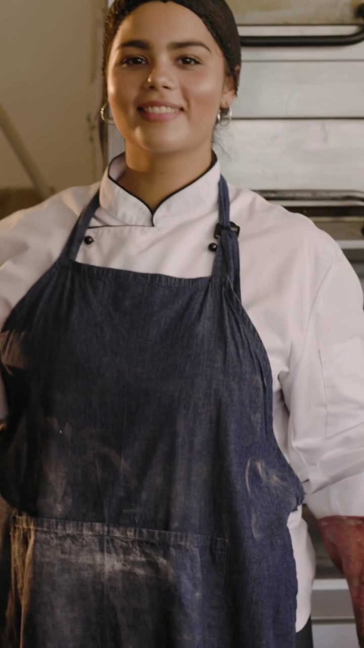 Video vertical: Chef caminando desde la derecha, introduciendo una bandeja de masa en el horno para hornear, con chaqueta y guantes.