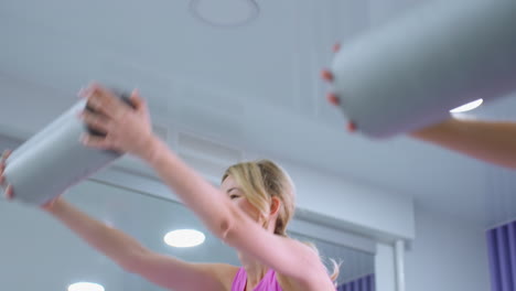 el traje de entrenamiento de la mujer levanta el rodillo de espuma por encima de la cabeza mientras sonríe y se estira hacia adelante, un gran espejo refleja el fondo del gimnasio, con otra persona realizando el mismo movimiento a un ritmo más rápido