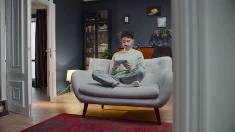 joven feliz sentado en el sofá con las piernas cruzadas y viendo algo divertido en el teléfono inteligente