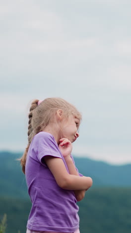 coquettish little child with blonde hair touches cheek by finger against distant mountains and grey sky at eco resort closeup slow motion