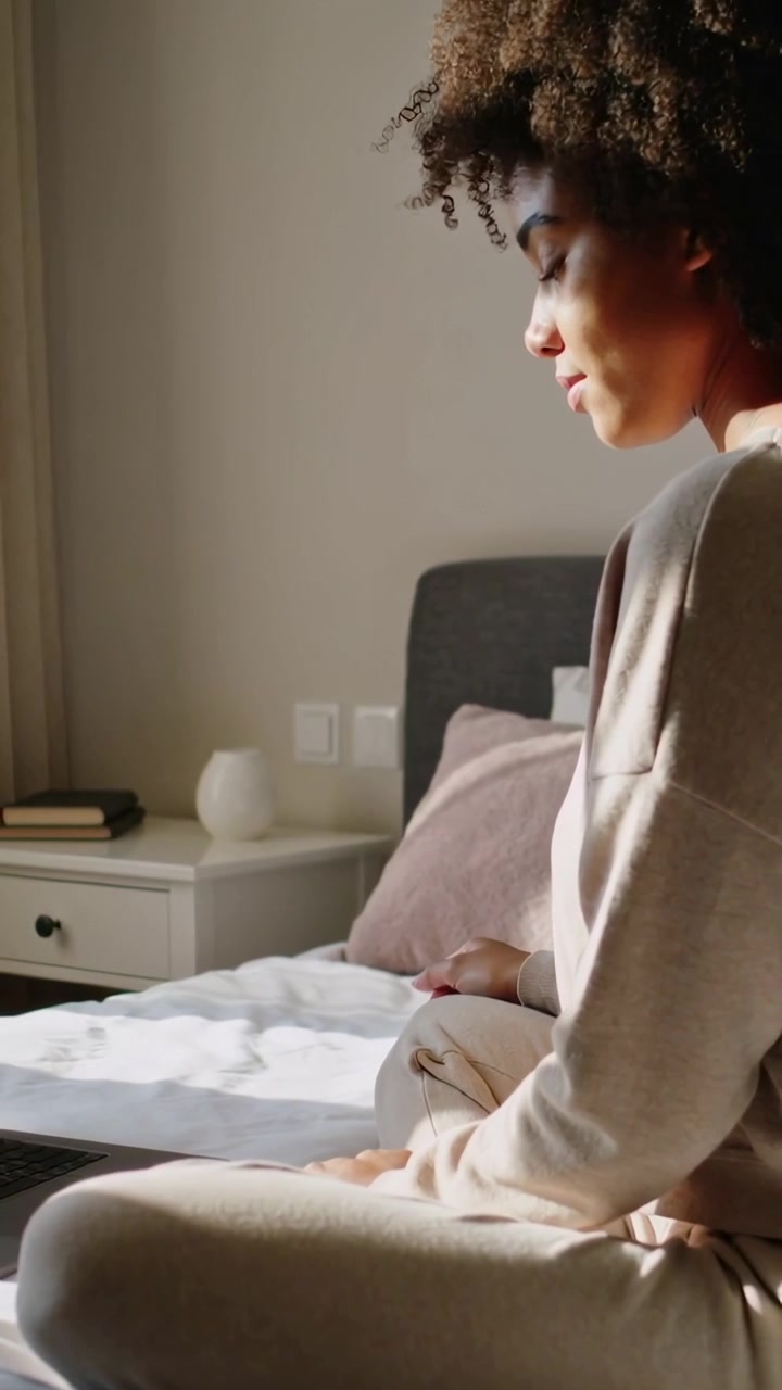 Vertical video: Shifting, woman in sweats responding to sun and watching laptop on bed, copy space