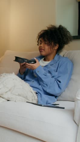 una mujer joven relajándose en un sofá, usando auriculares y un controlador de videojuegos.
