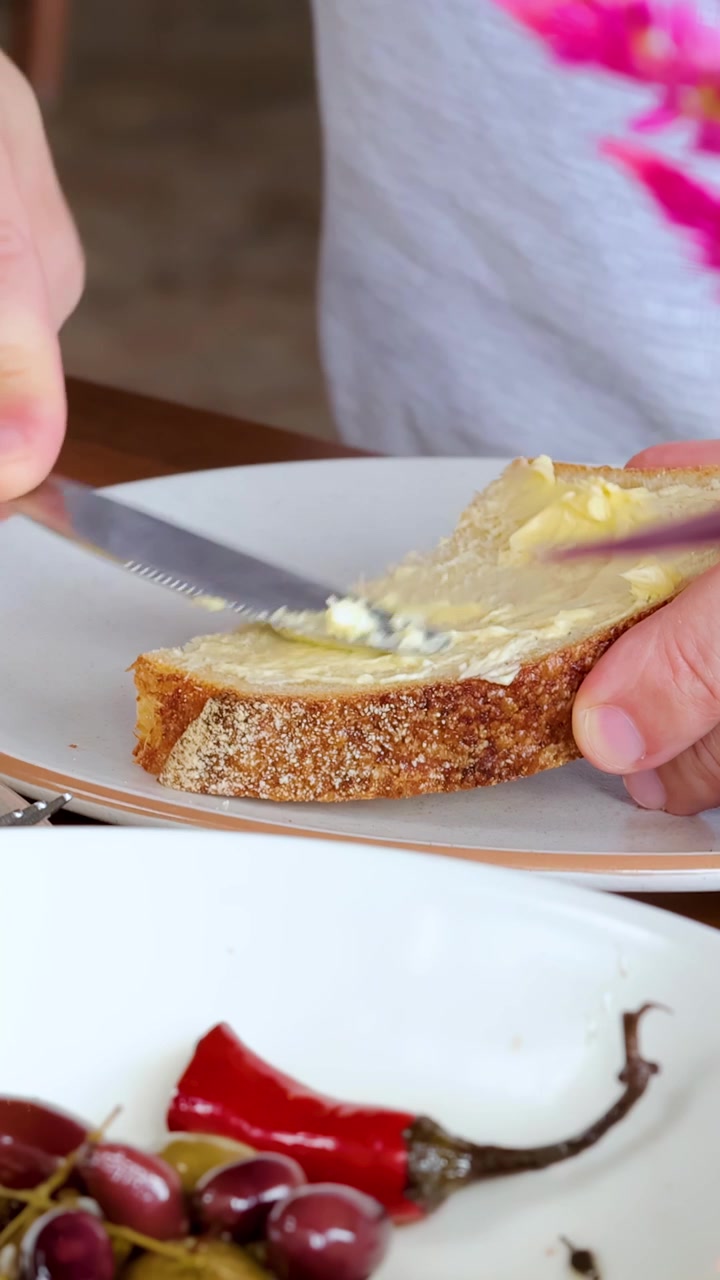 A person spreads butter onto a slice of rustic bread using a knife, with natural daylight and a breakfast plate of grapes and peppers nearby