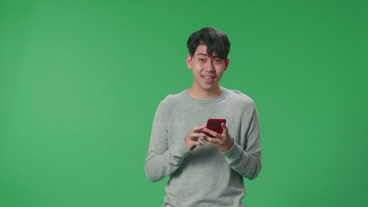 Smiling Young Asian Man Use Mobile Phone And Pointing To Smart Phone Screen And Positively Shaking His Head While Standing In The Green Screen Studio