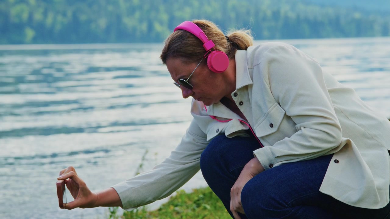 Woman wearing pink headphones crouches near water. She focuses on phone, possibly capturing nature. Scene feels calm and serene. Ideal for travel, wellness, or outdoor lifestyle projects