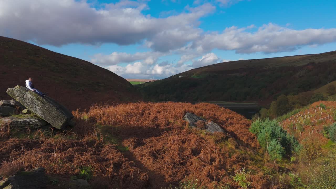 un joven encaramado en un afloramiento rocoso sustancial, perdido en la contemplación, ya que las imágenes aéreas giran graciosamente alrededor del páramo