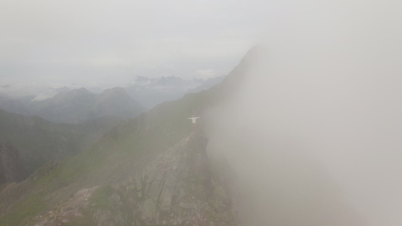 Revealing aerial around young hiker man standing on Mountain Peak Summit, moving through the clouds Fl&oslash;ya and Djevelporten above Svolv&aelig;r in Lofoten, Norway, aerial footage