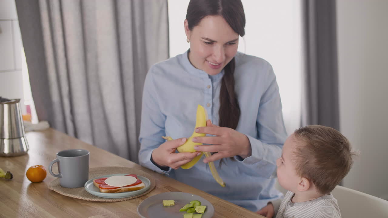 Mother Peeling Banana And Giving A Piece To His Cute Little Son Sitting In High Chair Next To Her At Home