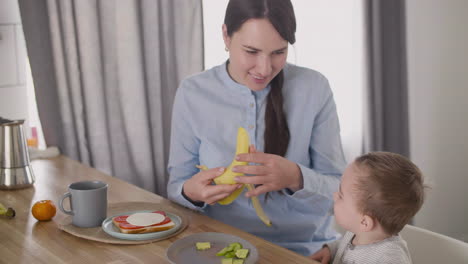 mother peeling banana and giving a piece to his cute little son sitting in high chair next to her at home