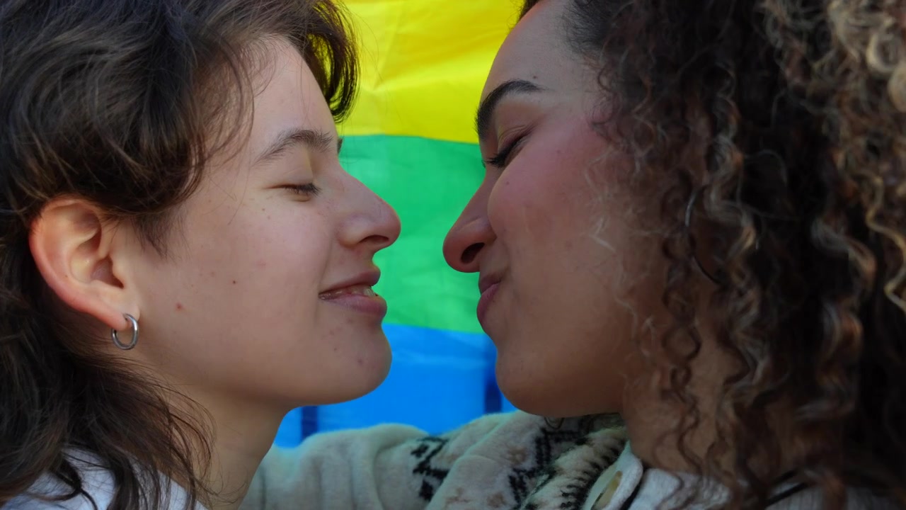 Young lesbian couple kissing and laughing together in front of a rainbow flag at a pride parade. Close up